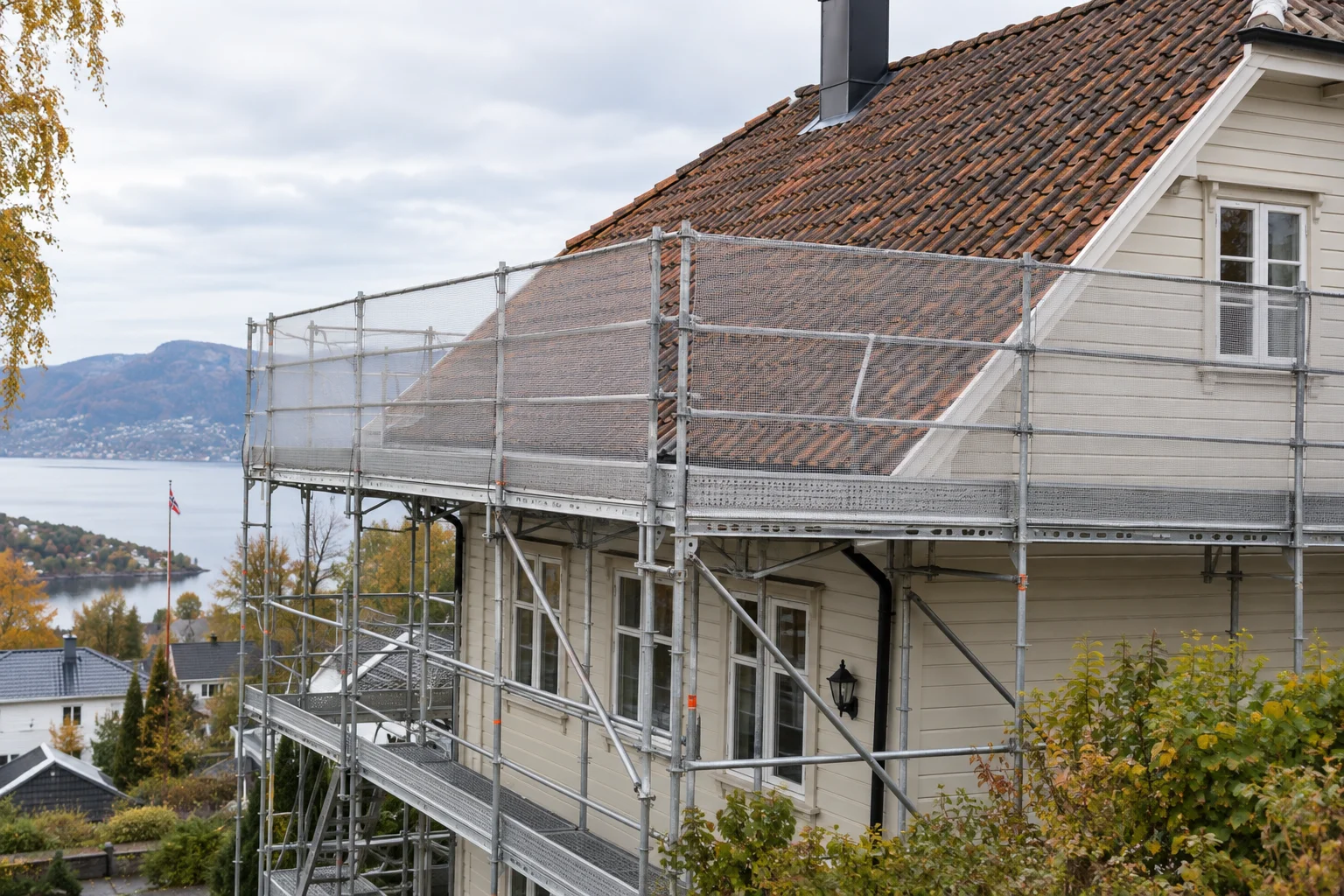 Stillas langs takfoten på et trehus med tegltak og netting ved øvre nivå.